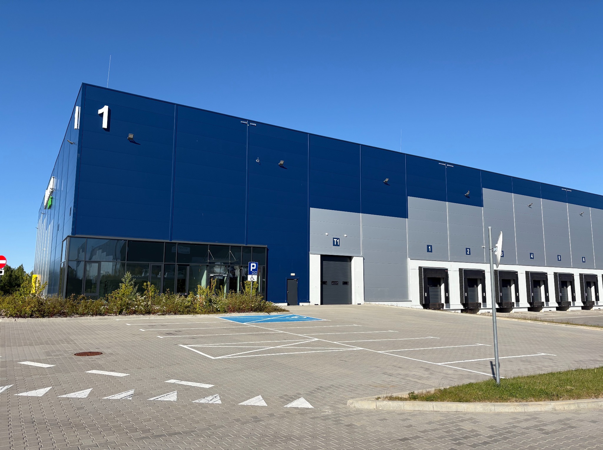Nowoczesny budynek magazynowy z elewacją w odcieniach niebieskiego i szarego, z rampami załadunkowymi i oknami. Widoczny parking z wyznaczonym miejscem dla niepełnosprawnych.