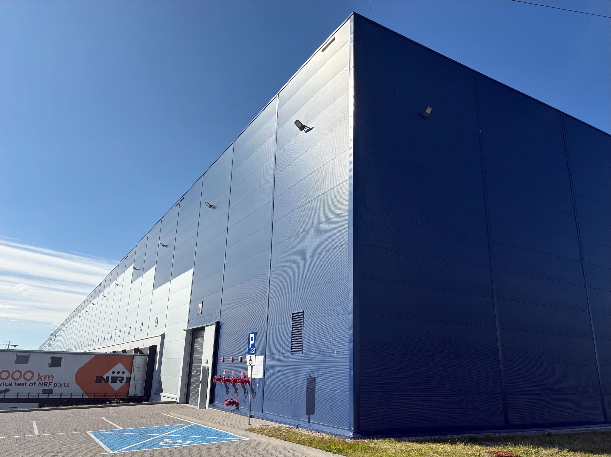 Narożnik hali magazynowej z płyt warstwowych w odcieniach szarości i granatu, oświetlonej promieniami słonecznymi. Widoczny fragment parkingu i instalacja przeciwpożarowa.