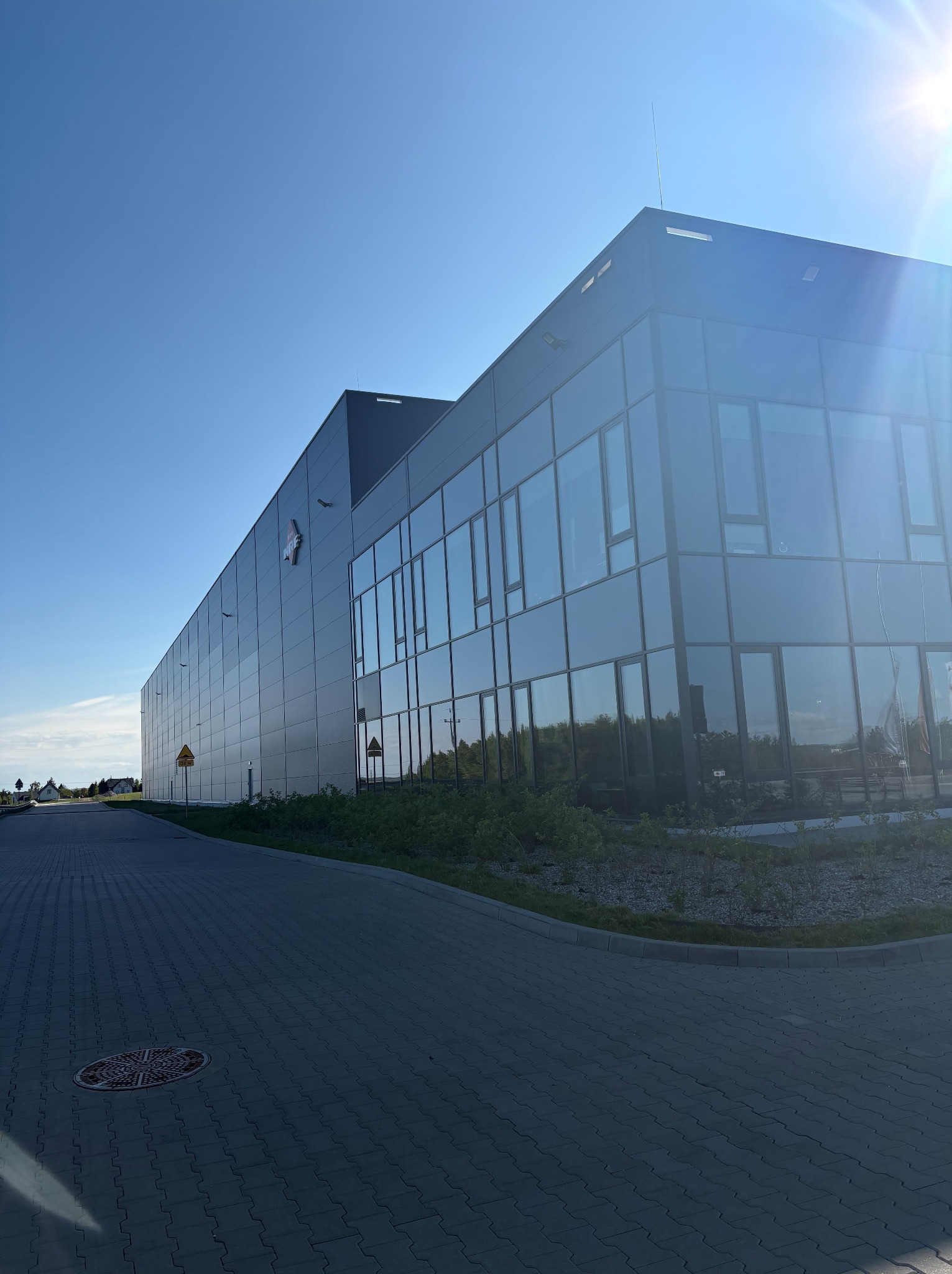 Nowoczesny budynek biurowo-magazynowy z szarymi panelami i szklaną fasadą, widoczny z poziomu chodnika w słoneczny dzień. Na elewacji logo firmy.