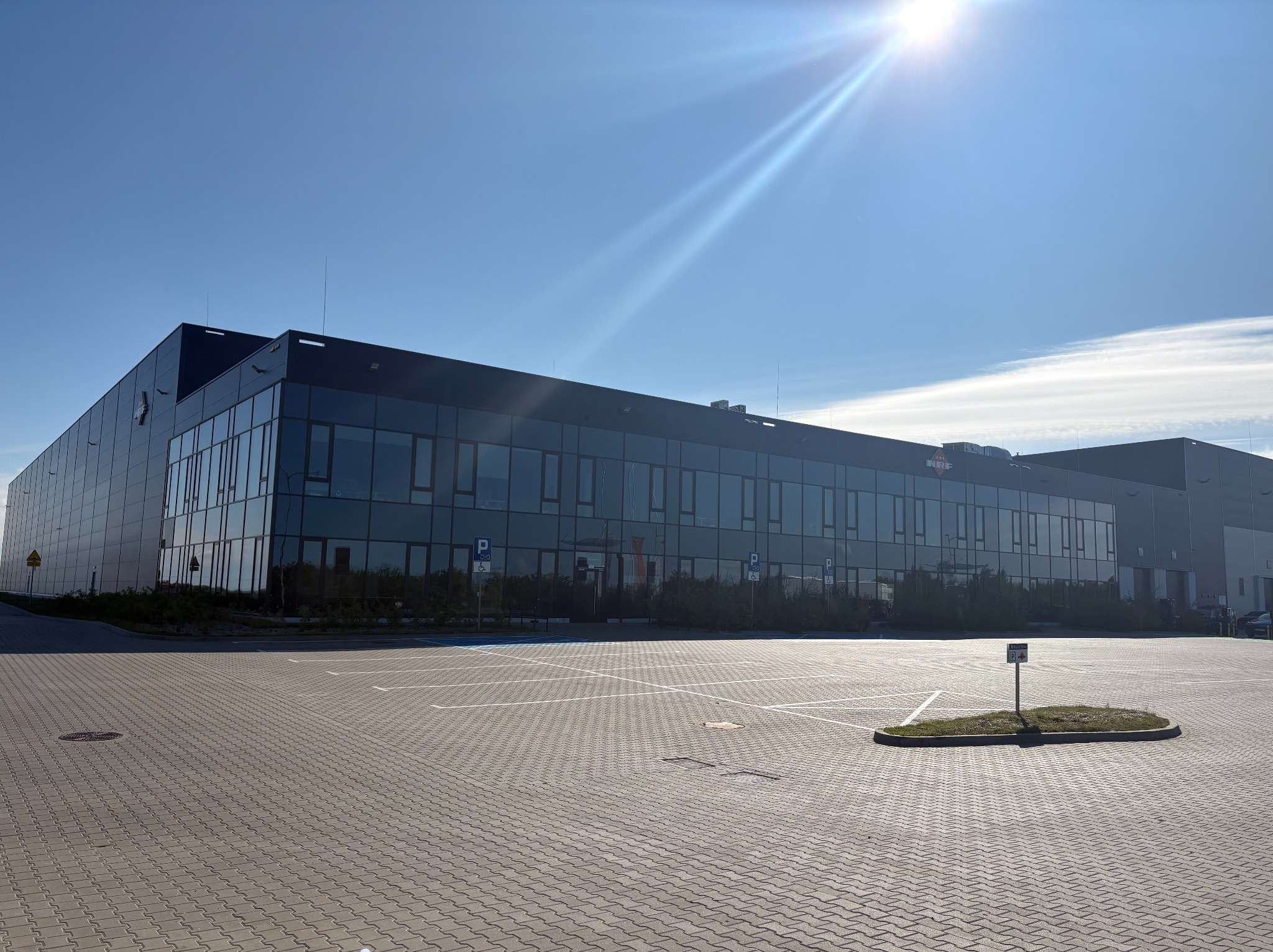 Nowoczesny budynek biurowy z ciemną elewacją i licznymi oknami, widoczny z rozległego parkingu w słoneczny dzień. Uwagę zwraca minimalistyczny design i symetryczny układ.