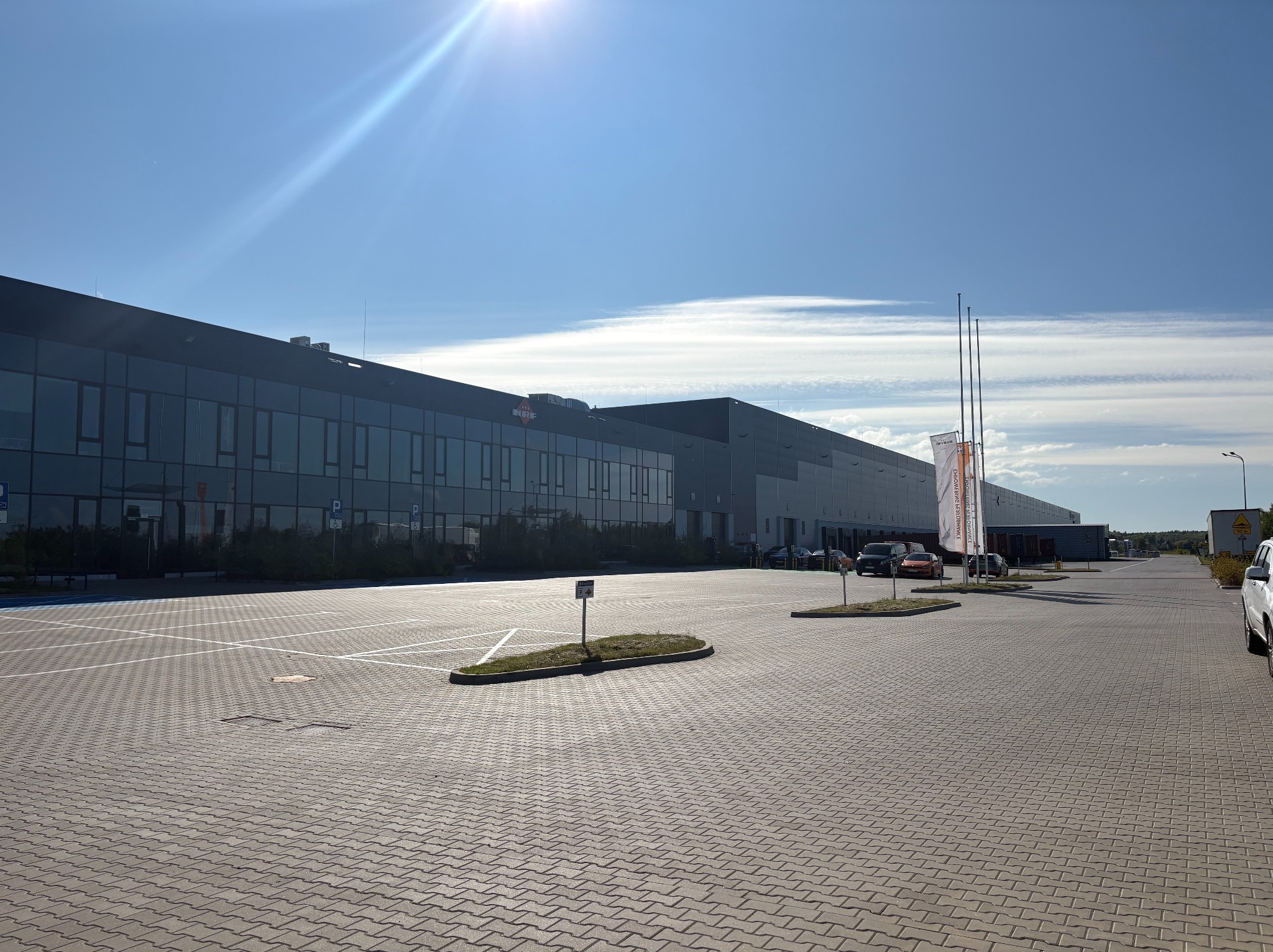 Nowoczesny budynek magazynowo-biurowy z ciemną elewacją, widok z parkingu w słoneczny dzień. Widoczne okna, logo firmy, flagi i zaparkowane samochody.