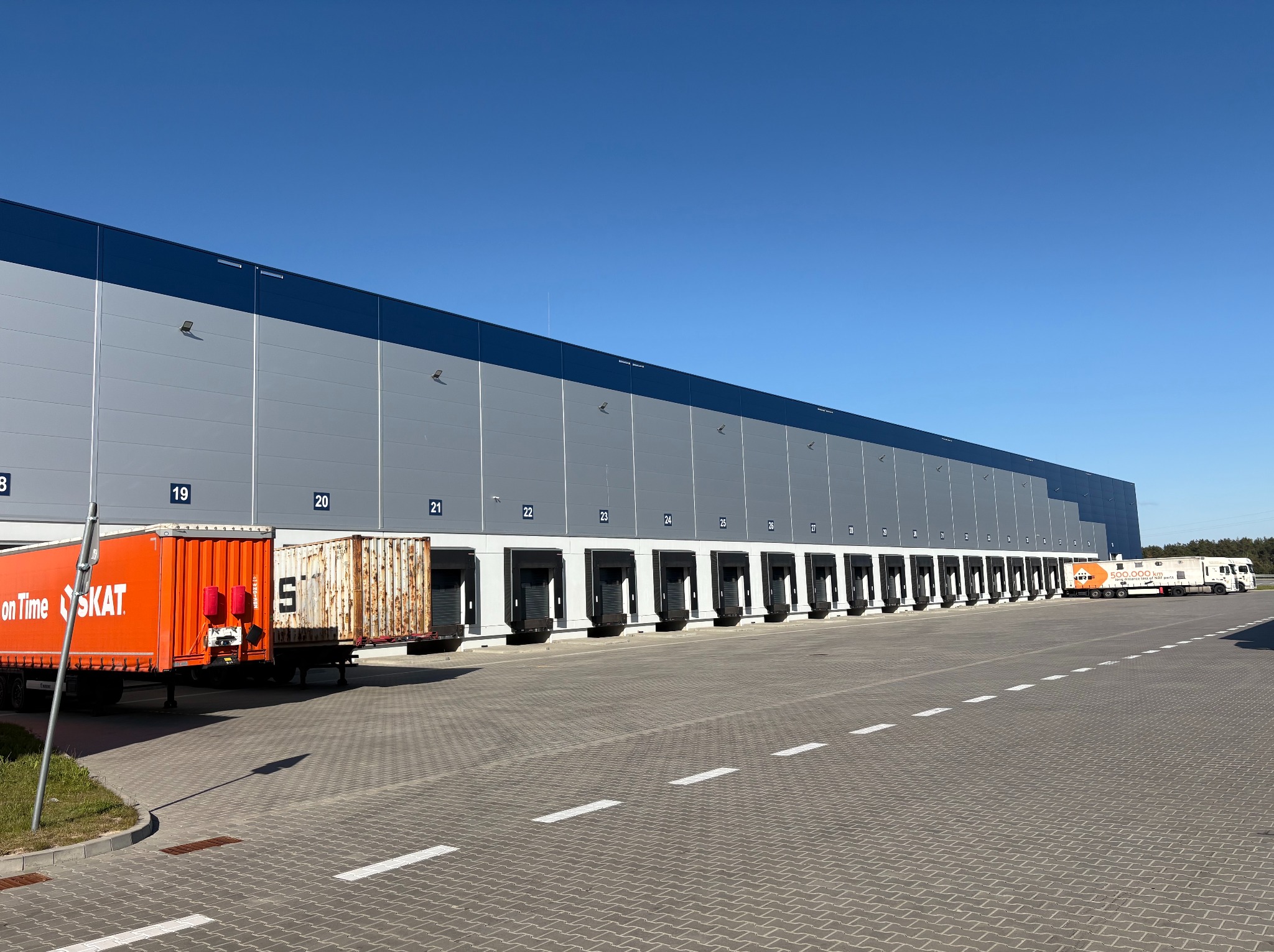 Długi budynek hali magazynowej z rampami załadunkowymi, widoczny z zewnątrz, z zaparkowanymi pojazdami ciężarowymi. Płyty warstwowe w szarym odcieniu.