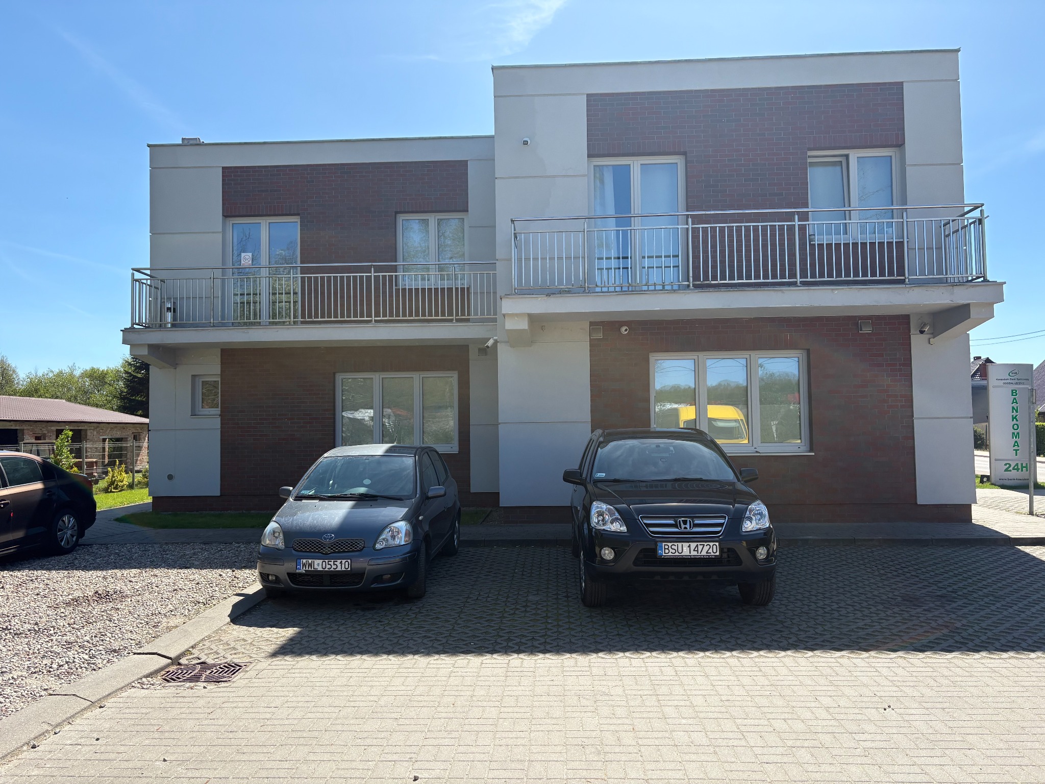 Dwupiętrowy budynek z ceglaną elewacją i balkonami, dwa samochody zaparkowane przed budynkiem na kostce brukowej, słoneczny dzień.