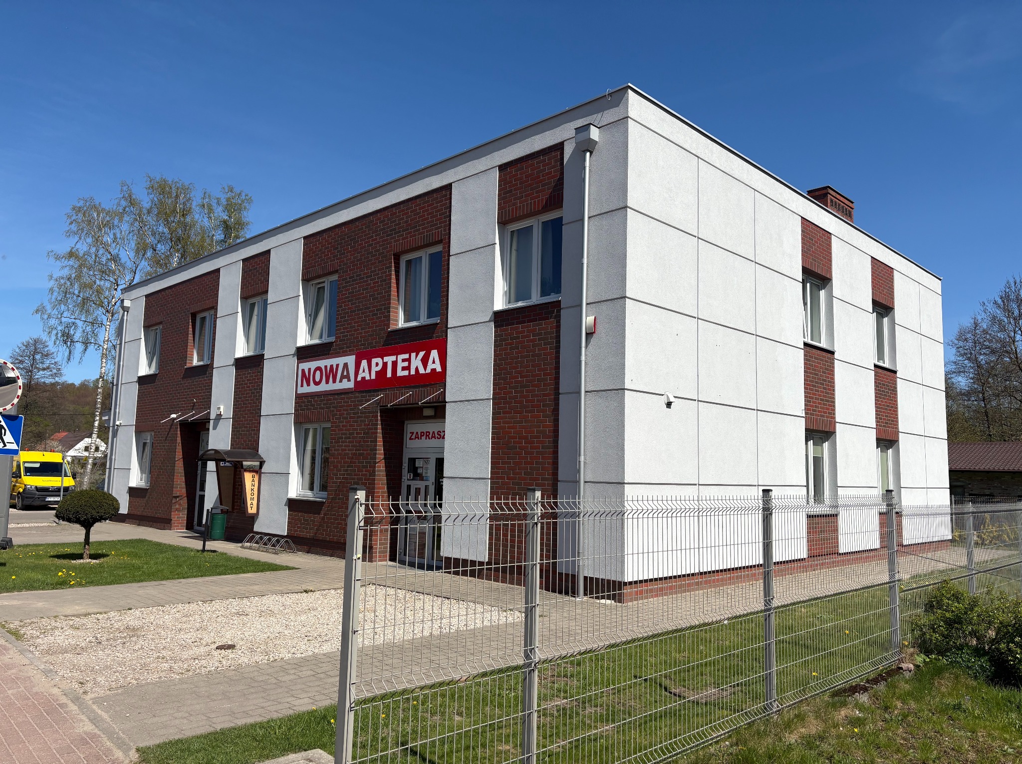 Dwukondygnacyjny budynek apteki z białymi panelami i ceglanymi wstawkami, widoczny z poziomu chodnika za metalowym ogrodzeniem. Błękitne niebo w tle.