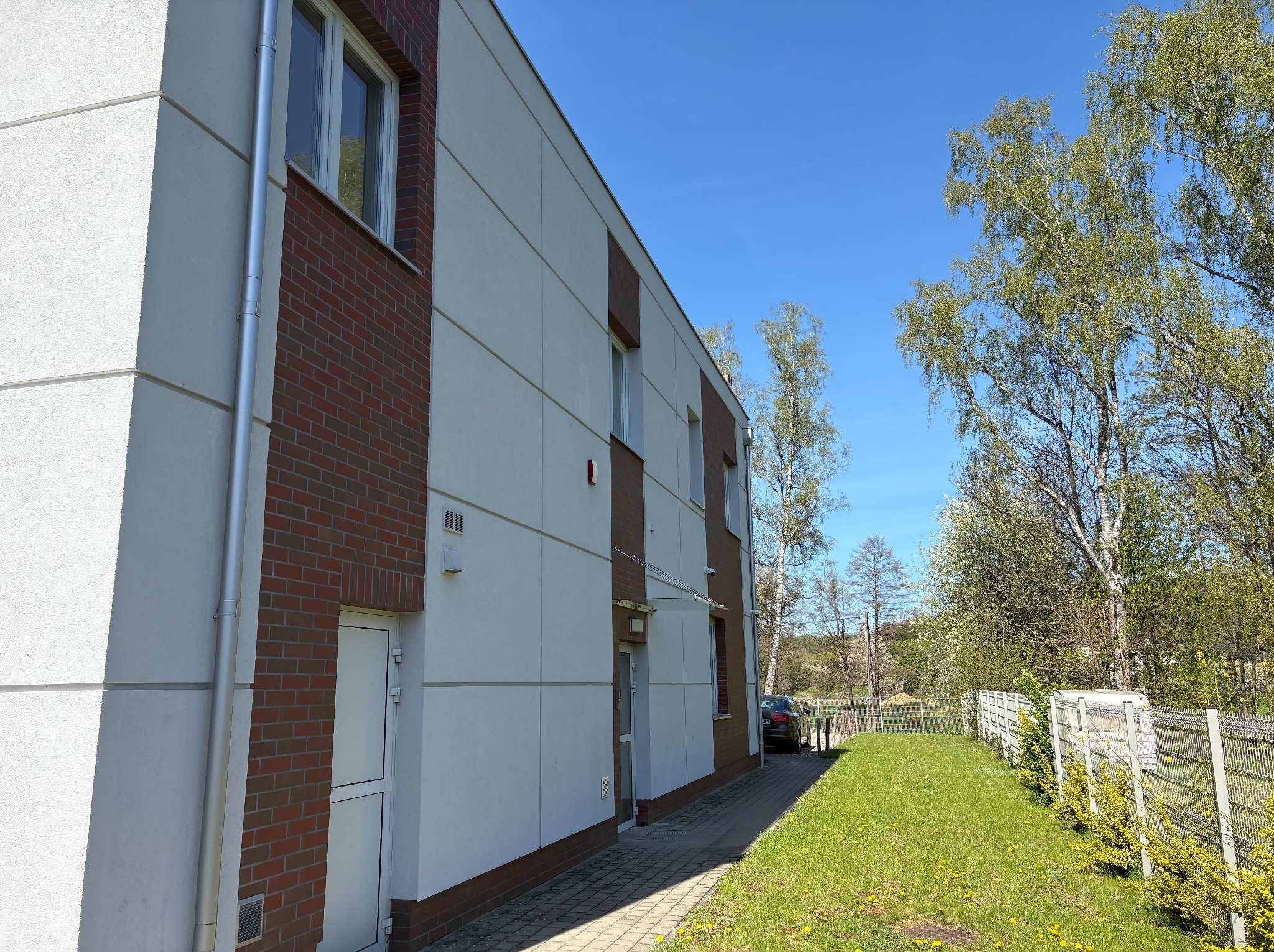 Dwukondygnacyjny budynek z elewacją w odcieniach bieli i cegły, z widocznymi oknami i rynnami, otoczony zielenią i ogrodzeniem.