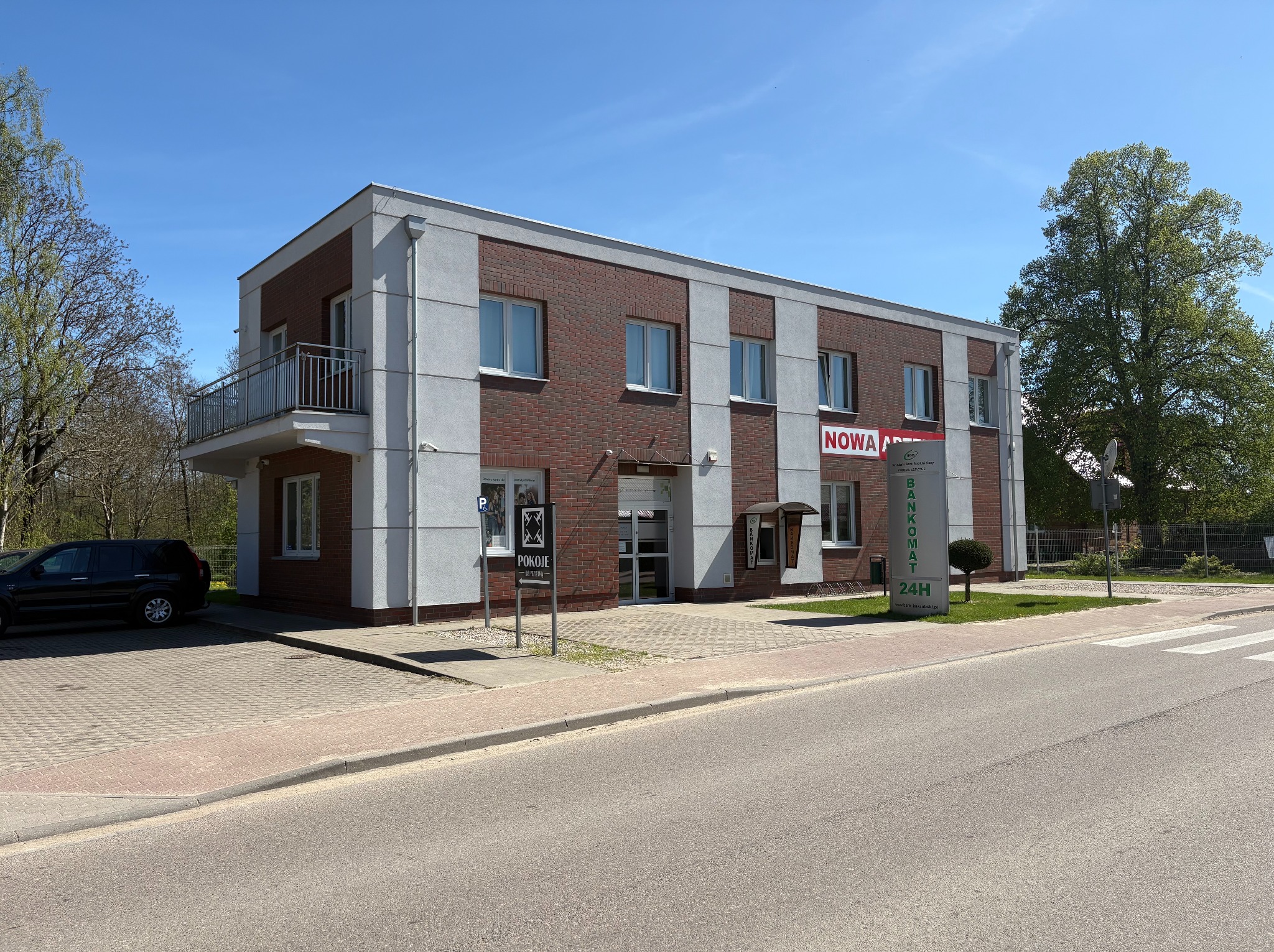 Dwukondygnacyjny budynek biurowy z balkonem, elewacją z czerwonej cegły i szarymi panelami. Widoczny szyld 'Bankomat 24H' i zaparkowany samochód.