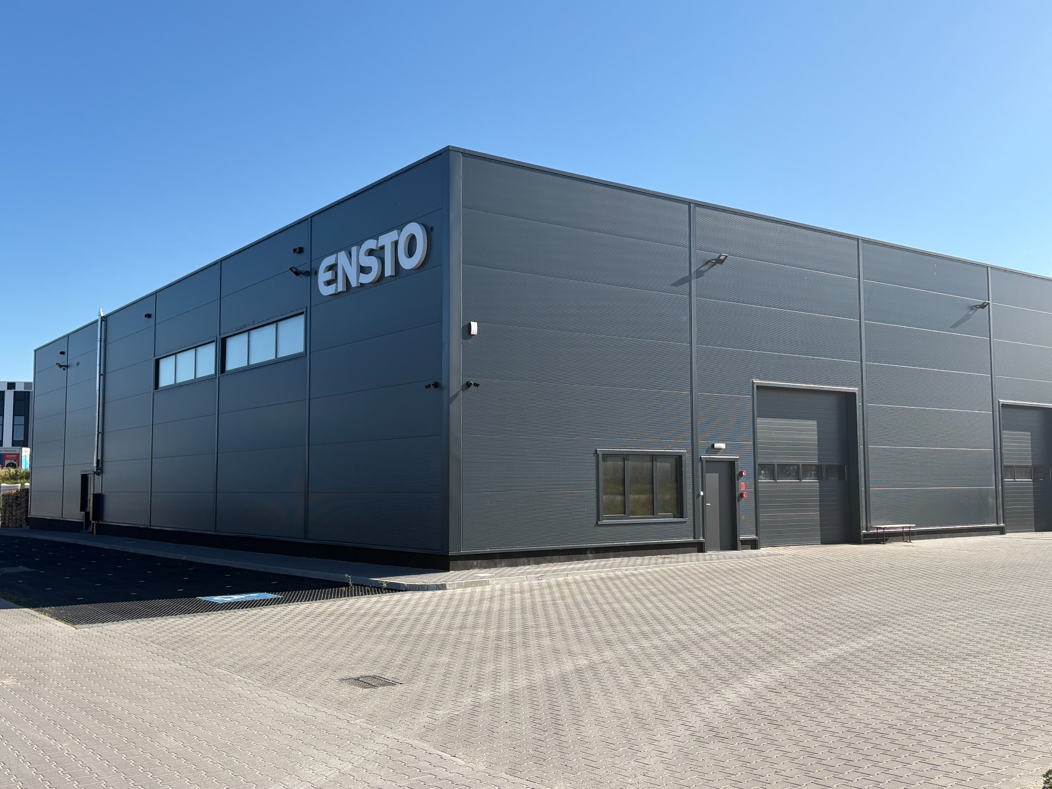 Nowoczesny, szary budynek przemysłowy z płyt warstwowych pod błękitnym niebem. Widoczne logo 'ENSTO' i dwie bramy wjazdowe.