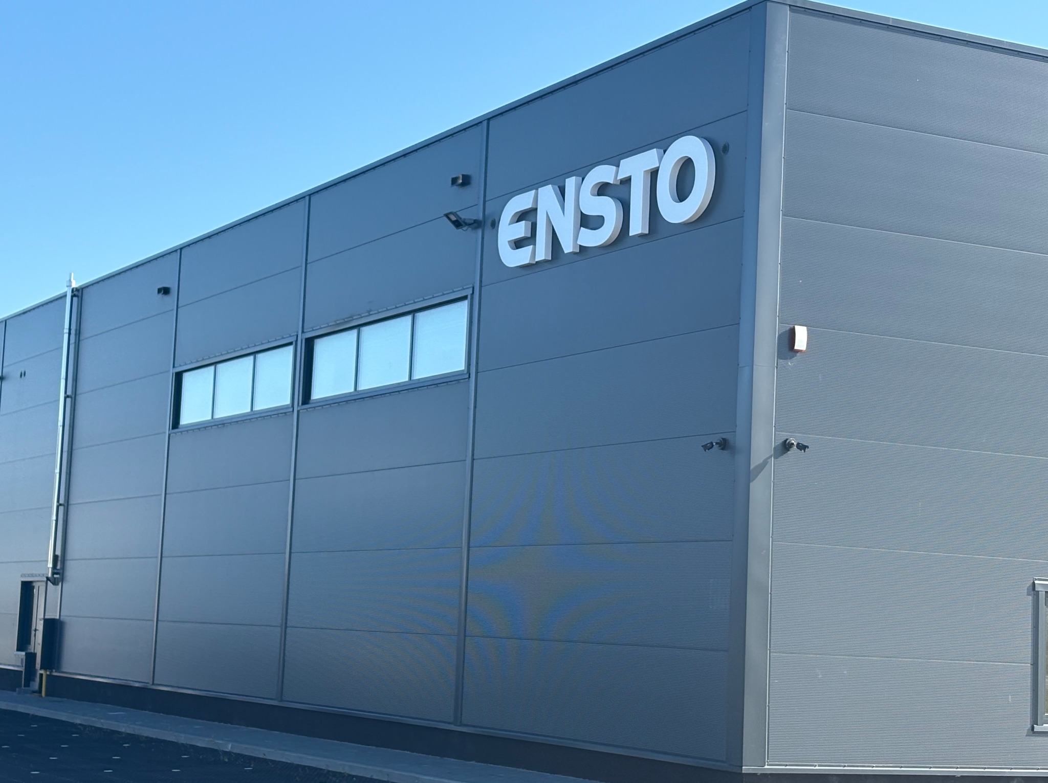 Szary budynek przemysłowy z płyt warstwowych, na którym widnieje białe logo firmy ENSTO. Widoczne okna i kamery przemysłowe. Błękitne niebo w tle.