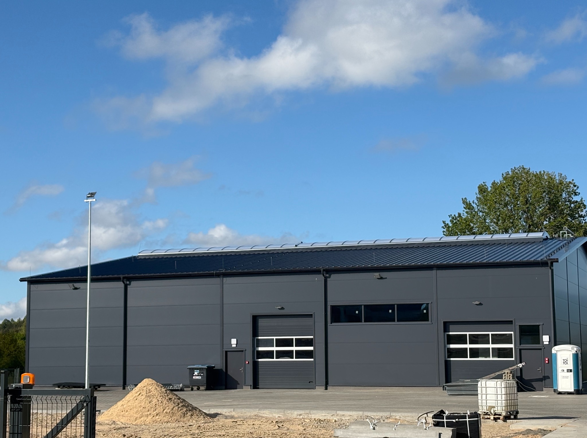 Nowoczesny budynek przemysłowy z grafitową elewacją, widoczne segmentowe bramy garażowe, okna, stos piasku i przenośna toaleta na placu budowy, błękitne niebo.