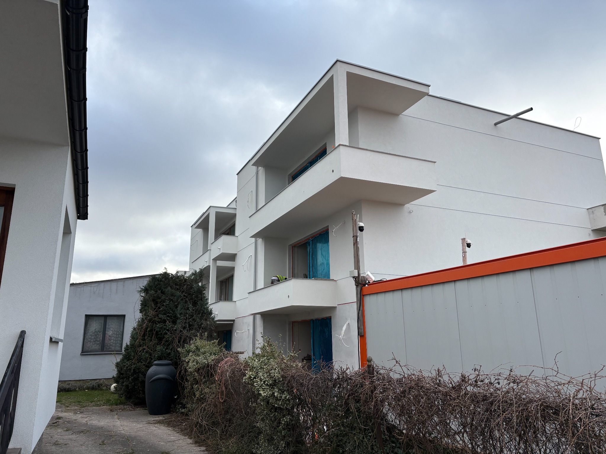 Nowoczesny, biały budynek wielorodzinny z balkonami, w trakcie wykończenia. Widoczne zabezpieczenia okien i kontener budowlany, sugerujące trwające prace.