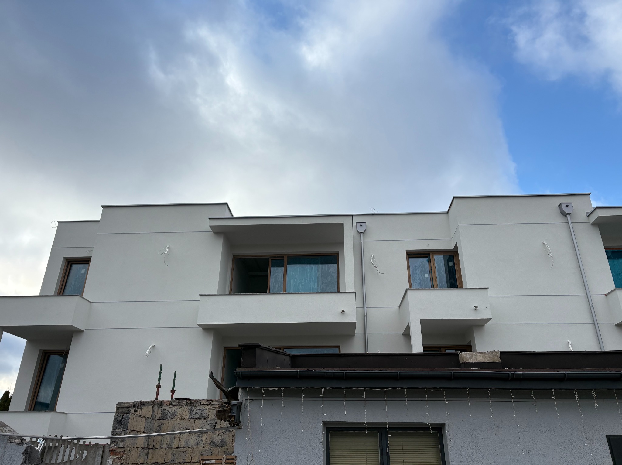 Nowoczesny, biały dom z balkonami i dużymi oknami, widoczny spod niższego budynku z elementami rozbiórki. Pochmurne niebo.