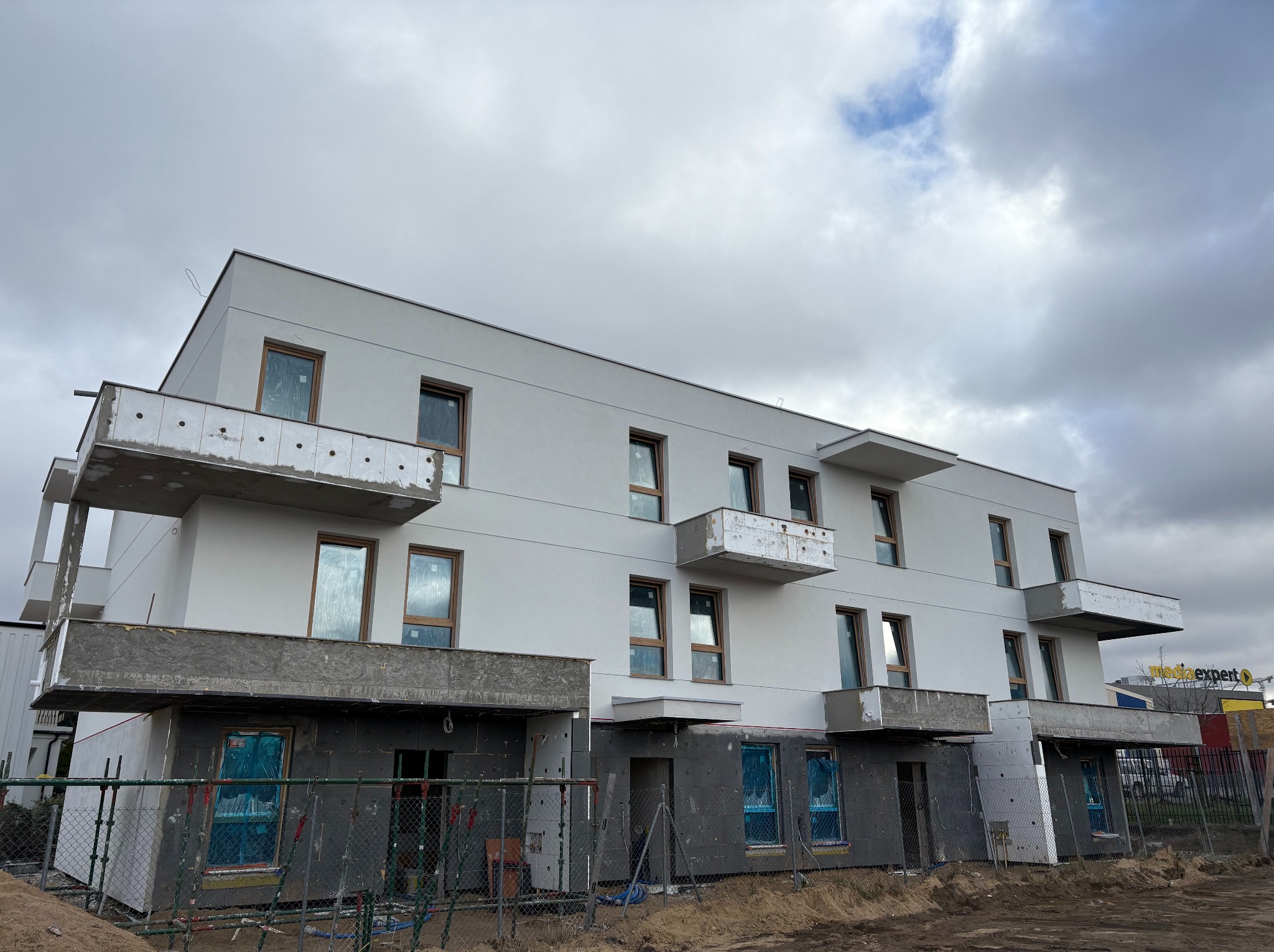 Wielopiętrowy budynek w trakcie budowy, z widocznymi balkonami i oknami zabezpieczonymi folią. Częściowo ocieplony, ogrodzony siatką, pochmurne niebo w tle.