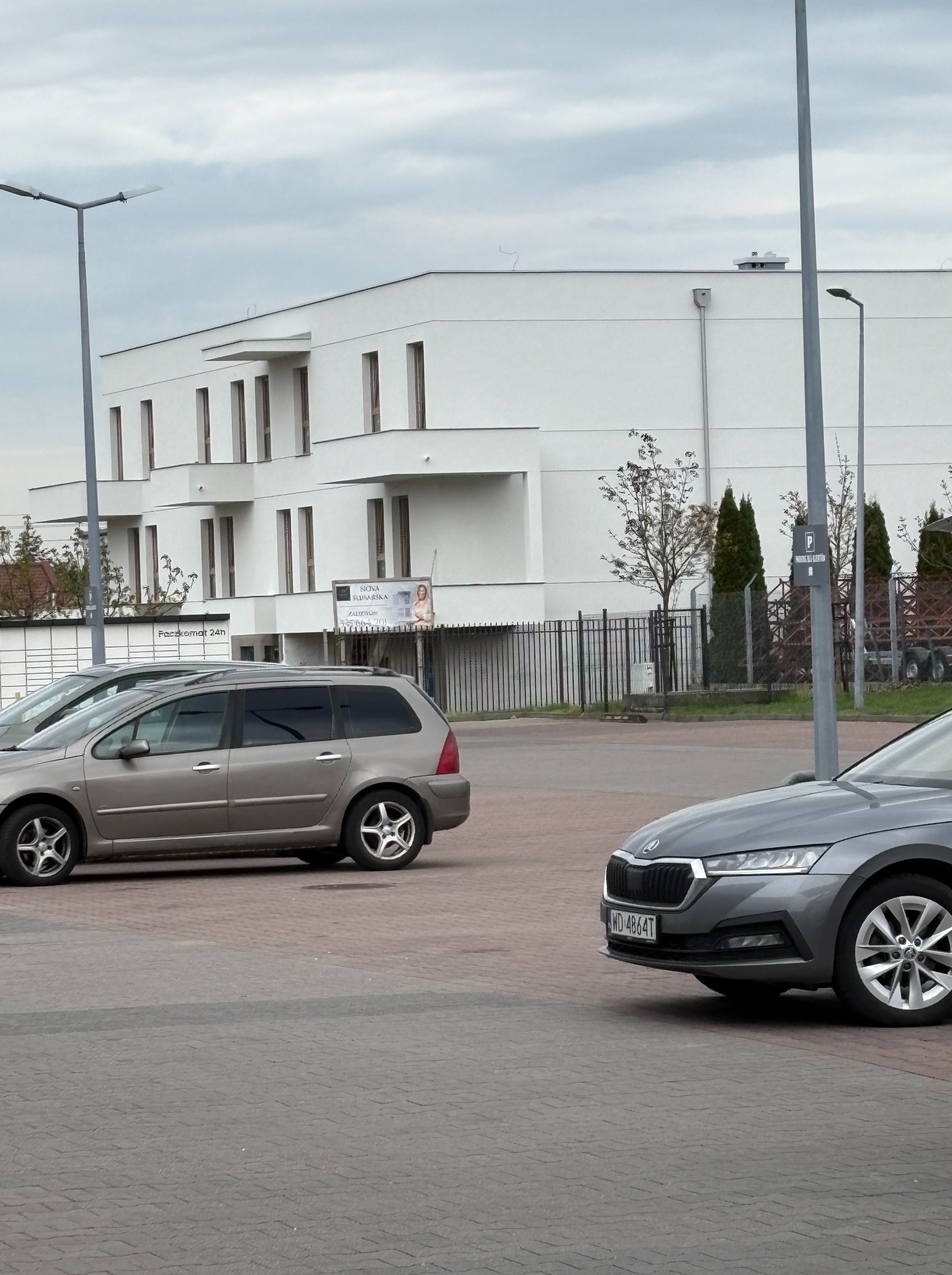 Nowoczesny, trzypiętrowy budynek z białymi elewacjami i regularnym układem okien, widziany z parkingu z dwoma samochodami w kadrze, przy pochmurnym niebie.