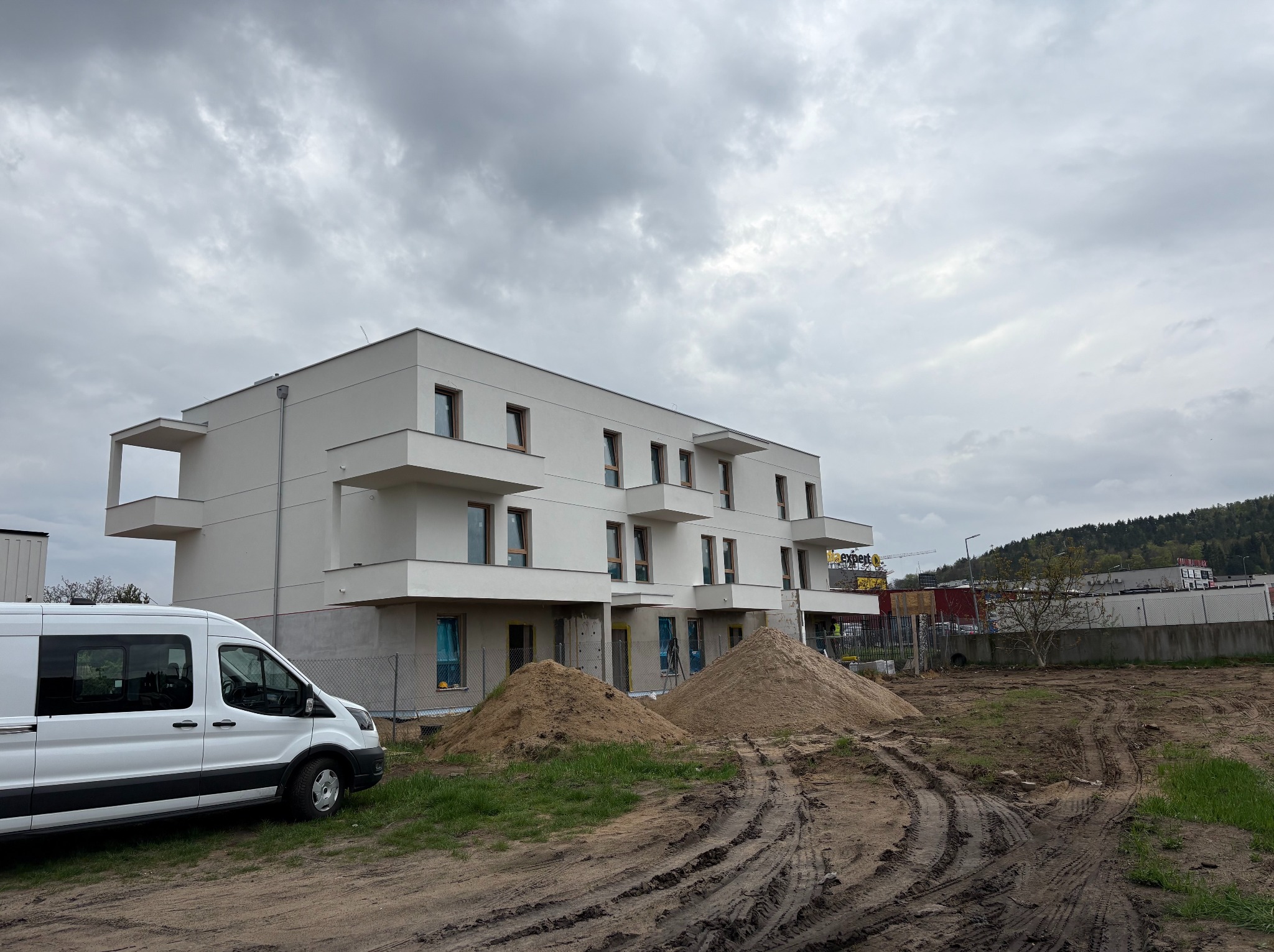 Nowoczesny, trzypiętrowy budynek w trakcie budowy, z widocznymi stertami piasku i białym vanem na pierwszym planie, na tle pochmurnego nieba.