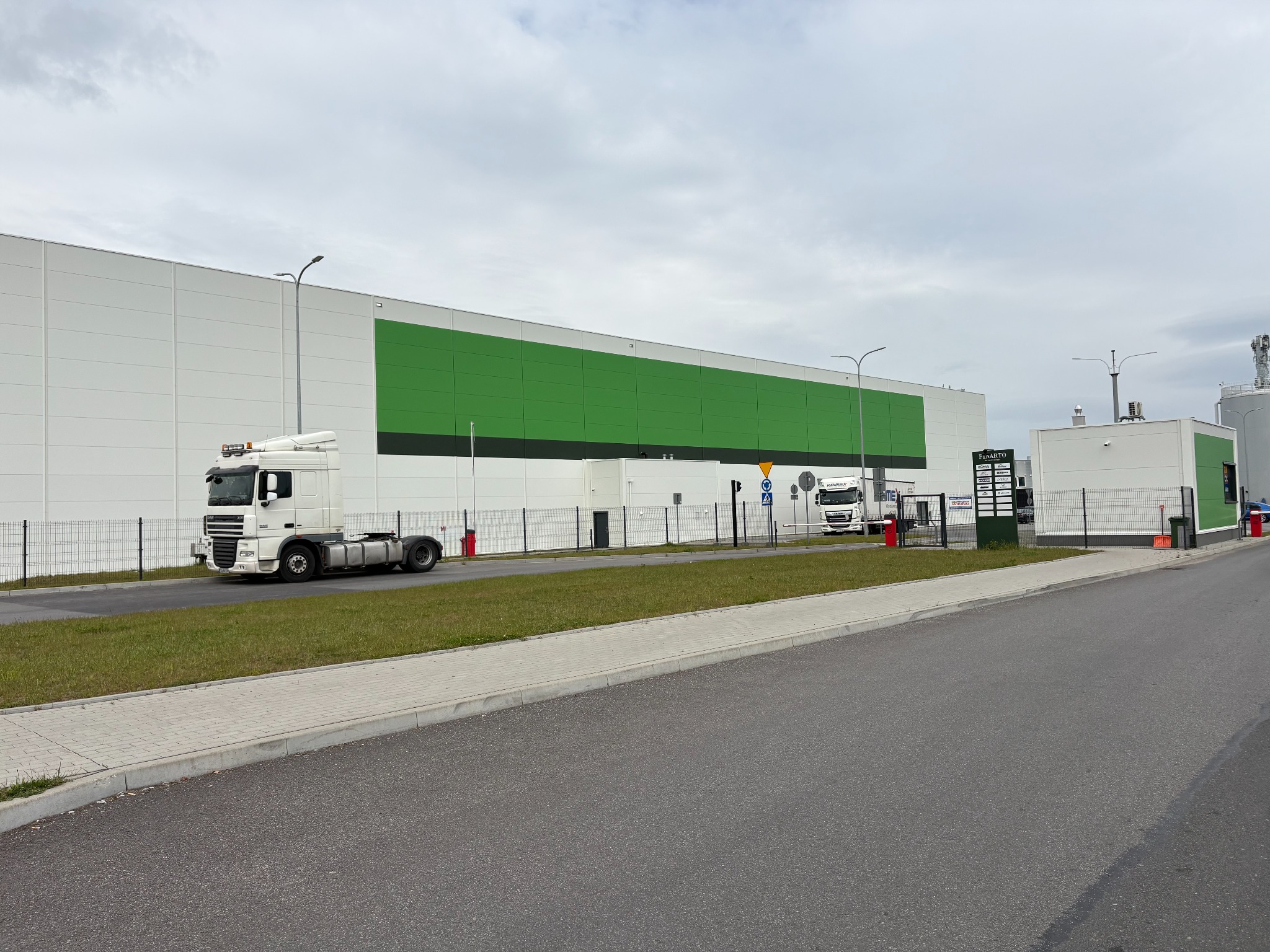 Biały budynek z zielonym pasem z płyt warstwowych, zaparkowany biały tir, brama wjazdowa, szara droga dojazdowa i chodnik.