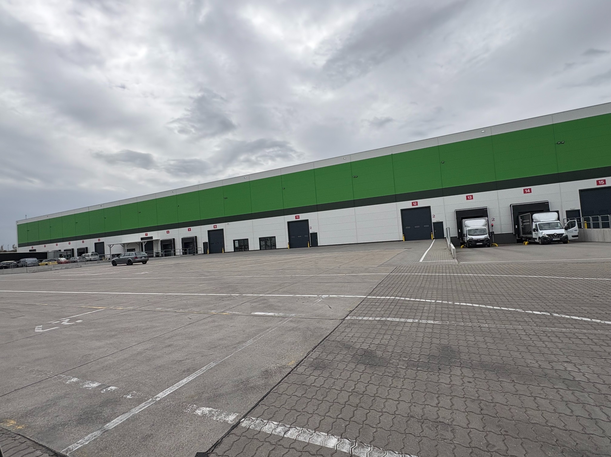 Hala magazynowa z zielonym pasem na górze, widoczne bramy załadunkowe z numerami, na placu manewrowym zaparkowane samochody dostawcze.