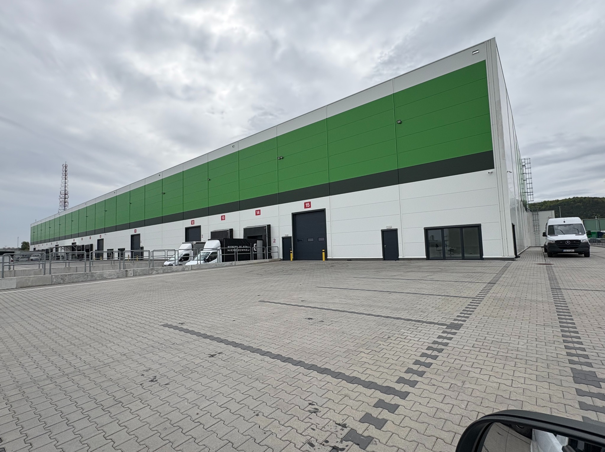 Nowoczesny budynek magazynowy z zielono-białą elewacją, widok z zewnątrz, z rampami załadunkowymi i dwoma białymi samochodami dostawczymi na brukowanym placu.
