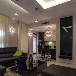 Nowoczesny salon z czarną skórzaną sofą narożną, stolikiem kawowym z czarnego szkła, kremowymi zasłonami i widokiem na kuchnię z kryształowym żyrandolem.