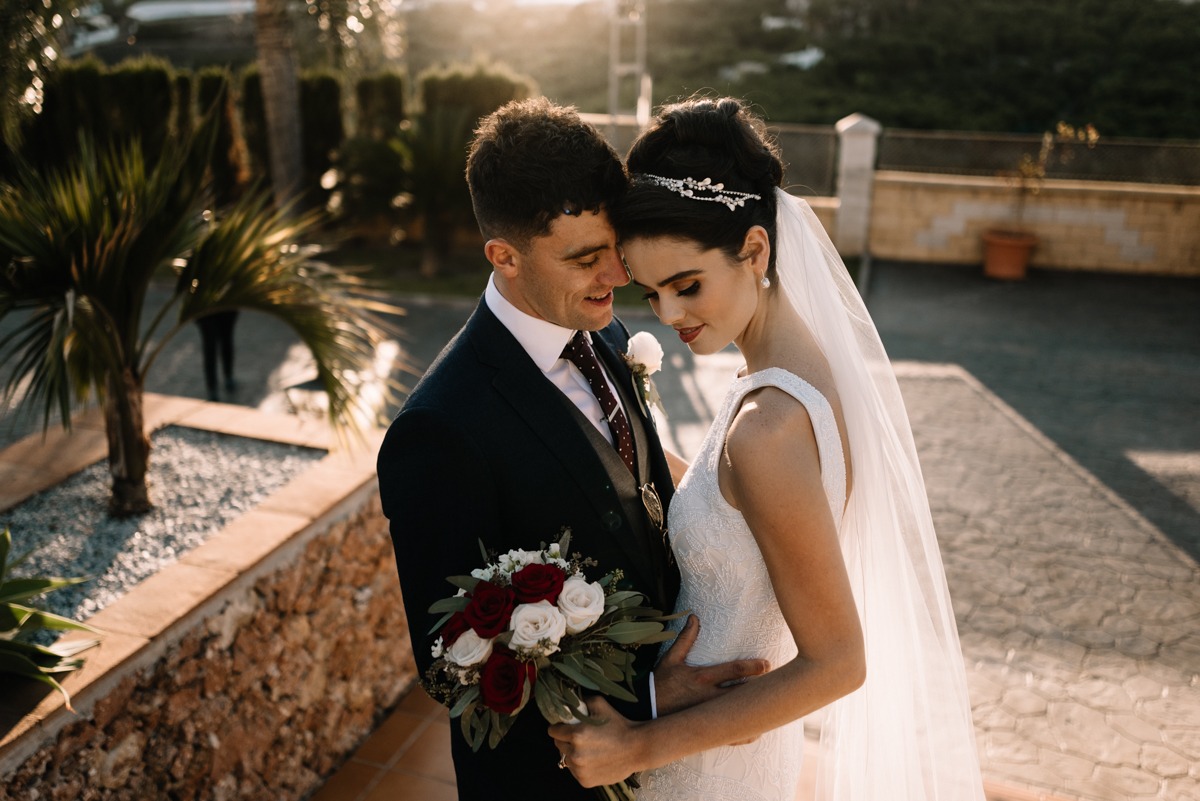 Panna młoda w sukni z koronkowymi aplikacjami i welonem, trzyma bukiet z czerwonych i białych róż, wtulona w pana młodego w ciemnym garniturze z butonierką, na tle śródziemnomorskiej roślinności...