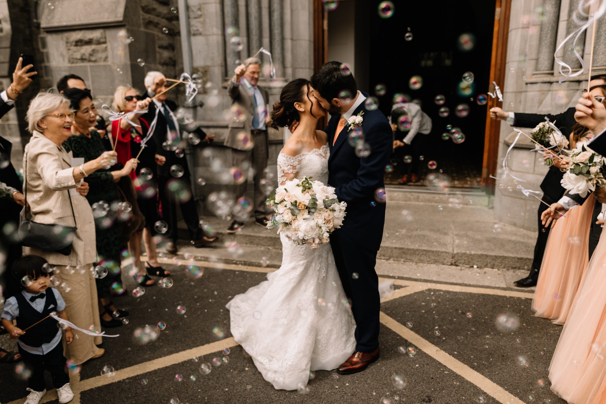 Pocałunek pary młodej na tle gości wypuszczających bańki mydlane przed kamiennym budynkiem, mały chłopiec w garniturze z boku.