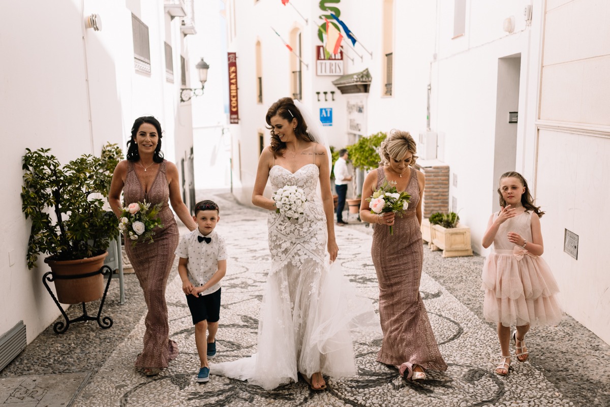 Panna młoda w białej sukni ślubnej, dwie druhny w długich sukniach, chłopiec z muszką i dziewczynka w tiulowej sukience idą uliczką w hiszpańskim miasteczku.