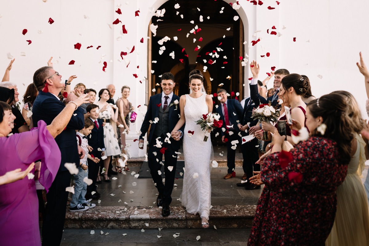 Para młoda wychodzi z kościoła podczas ceremonii ślubnej, obsypywana płatkami róż, goście weselni w tle.