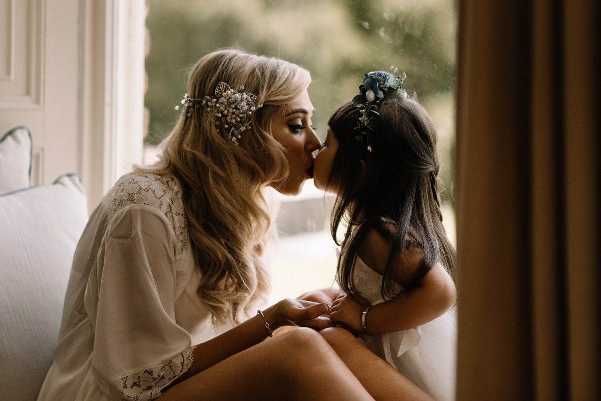 Kobieta w szlafroku z koronkowymi rękawami i ozdobą we włosach całuje w usta małą dziewczynkę z wiankiem na głowie, siedzącą na parapecie okna.