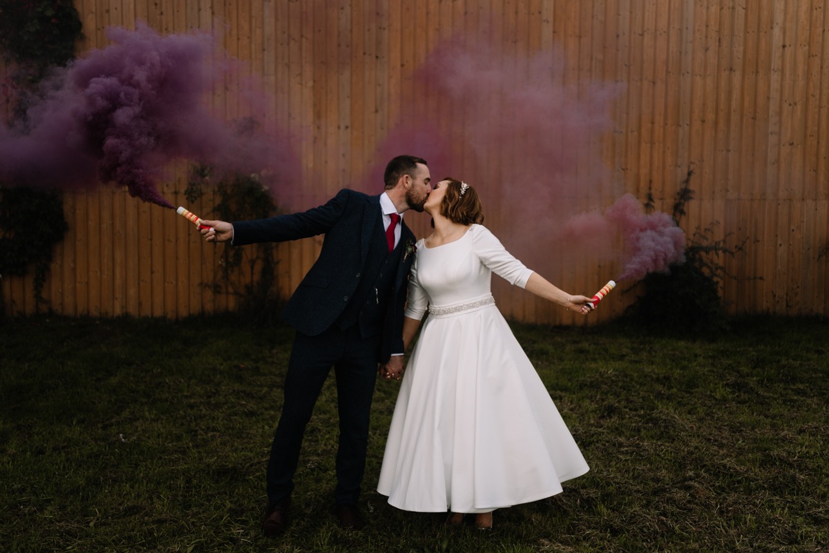Pocałunek nowożeńców na tle drewnianego płotu z purpurowymi dymami z rac.