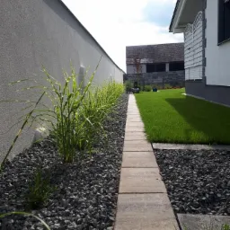 Ścieżka z płyt betonowych wzdłuż szarej ściany i trawnika, obsadzona ozdobną trawą i wysypana szarym grysem, w tle budynek mieszkalny.