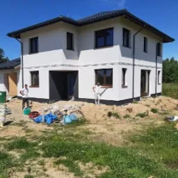 Dwukondygnacyjny dom jednorodzinny z białą elewacją, częściowo pomalowany na czarno u dołu, widoczni dwaj pracownicy w kombinezonach, jeden oparty o samochód dostawczy, drugi stojący przy ścianie...