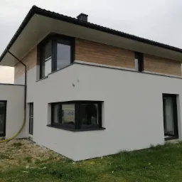 Nowoczesny dom jednorodzinny z jasną elewacją i elementami dekoracyjnymi imitującymi drewno na górnej części, czarne ramy okienne, widoczny fragment trawnika i żółty wąż przy ścianie