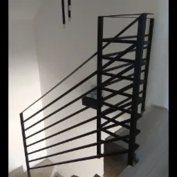 Nowoczesne schody metalowe z czarną balustradą o minimalistycznym designie, widoczne surowe betonowe stopnie i fragment wykończonej podłogi na wyższym poziomie.