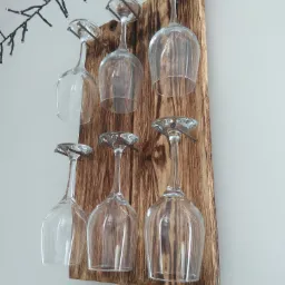 Drewniany wieszak na kieliszki do wina, wykonany z opalanej deski, z sześcioma kieliszkami zawieszonymi do góry dnem na gwoździach, ozdobiony lampkami choinkowymi w tle.