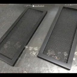 Dwa prostokątne, czarne, metalowe panele z siatką na szorstkiej, metalowej powierzchni roboczej, prawdopodobnie fragmenty większej konstrukcji.