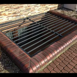 Zabezpieczenie okna piwnicznego z metalową kratą, obramowane ceglanym krawężnikiem, widoczne z poziomu chodnika z kostki brukowej.