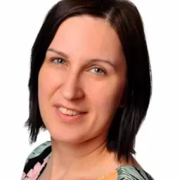 Portret uśmiechniętej kobiety z krótkimi, ciemnymi włosami i jasnymi oczami, ubrana w bluzkę z kwiatowym wzorem na białym tle.