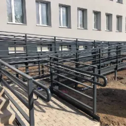 Zewnętrzne metalowe poręcze i podjazdy dla osób niepełnosprawnych przy budynku z szarą fasadą i rzędami okien, widoczne ślady prac ziemnych.