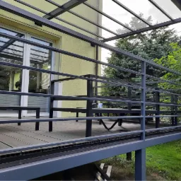 Zadaszony taras z metalową konstrukcją i balustradą w kolorze grafitowym, widok z zewnątrz, drewniana podłoga tarasu.