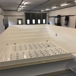 Wnętrze nowo wyprodukowanego, białego basenu z widocznymi schodami i zabezpieczającą folią na dnie, stojącego w hali produkcyjnej z oknami i bramą garażową w tle.