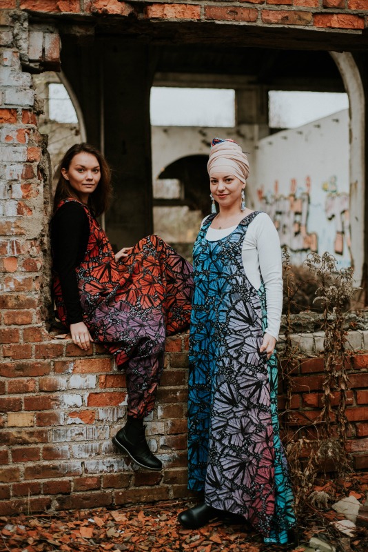 Dwie kobiety w sukienkach z motywem motyli, pozujące na tle zrujnowanego budynku z cegły.
