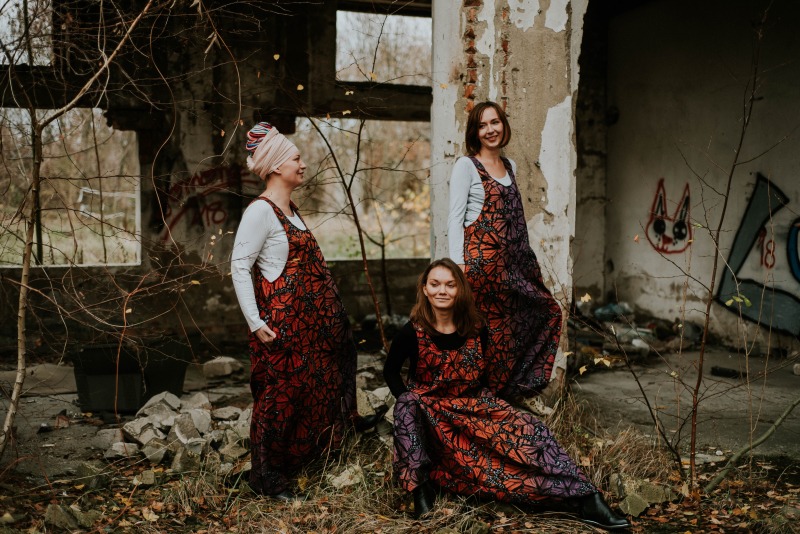 Trzy kobiety prezentują ręcznie szyte, długie ogrodniczki w kolorach jesieni z motywem geometrycznym na tle opuszczonego budynku z graffiti.