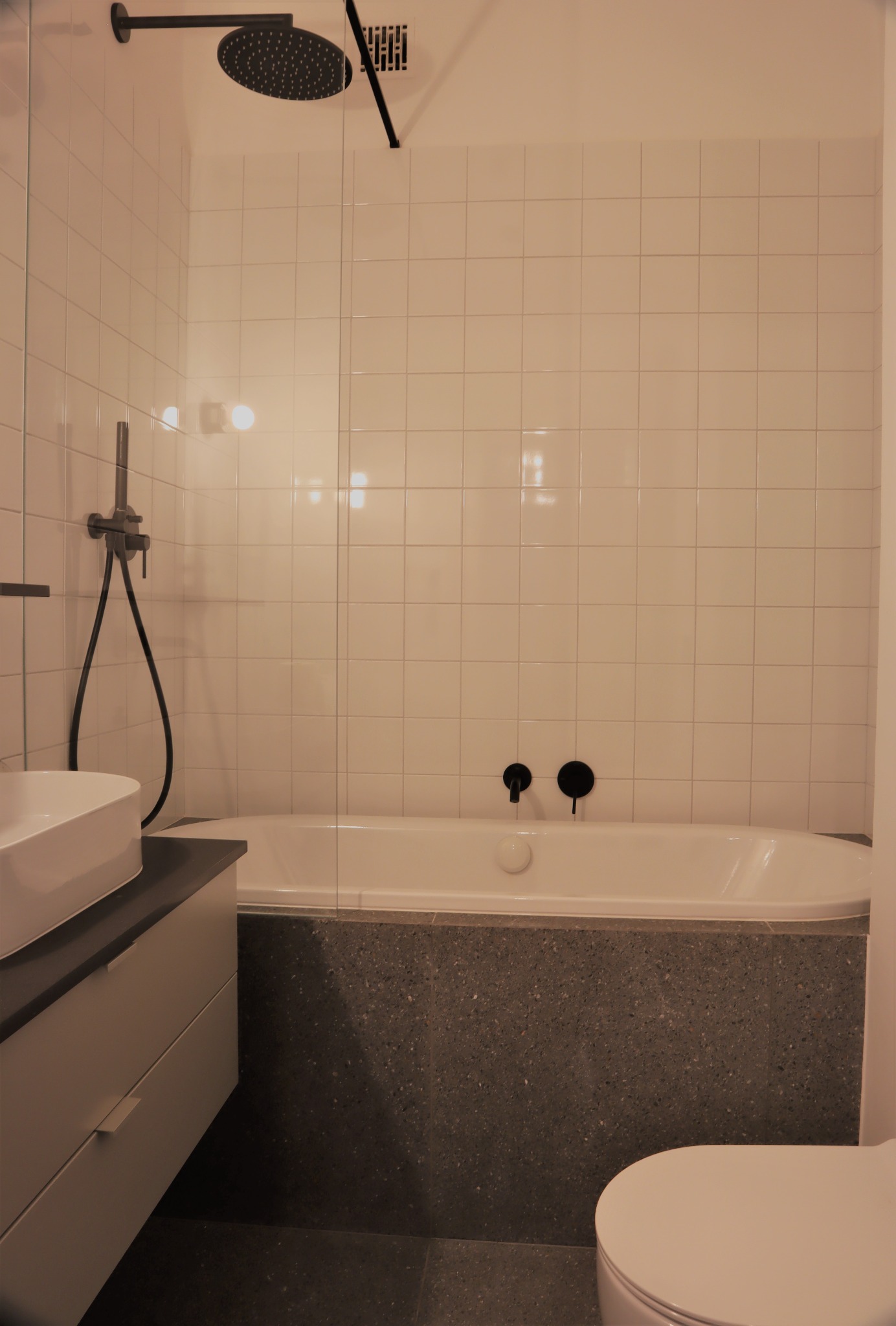 Wykończona łazienka z wanną obudowaną szarą mozaiką, białe płytki na ścianach, czarna armatura i deszczownica, umywalka nablatowa na szafce z szufladami, widoczny fragment toalety.