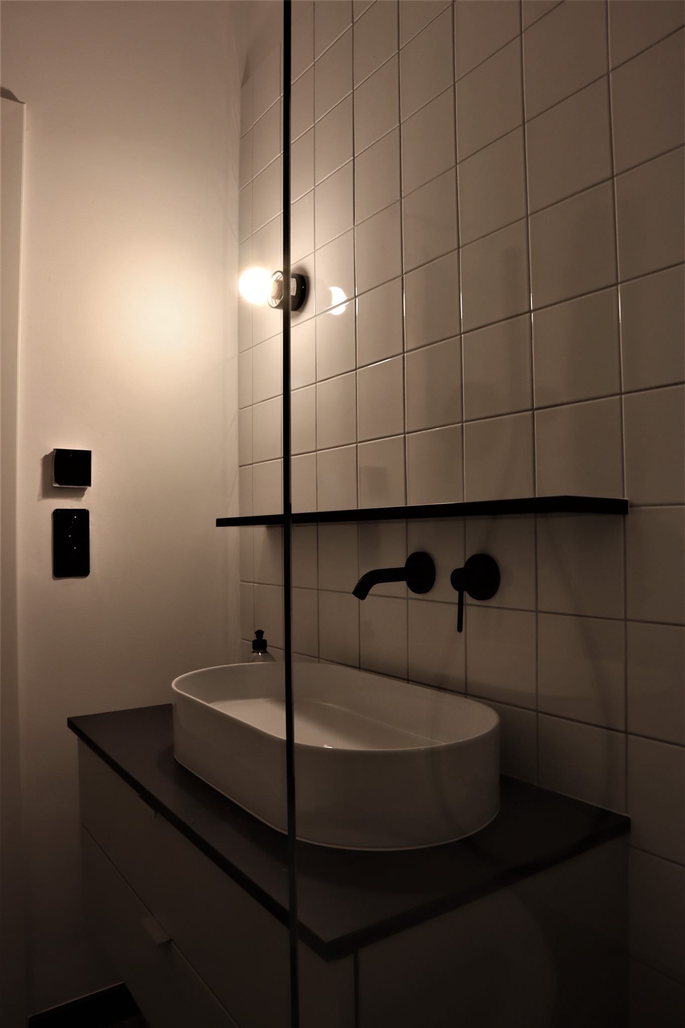 Nowoczesna łazienka z białą, owalną umywalką nablatową, czarną armaturą i płytkami w jasnym odcieniu, oświetlona pojedynczą żarówką bez klosza.
