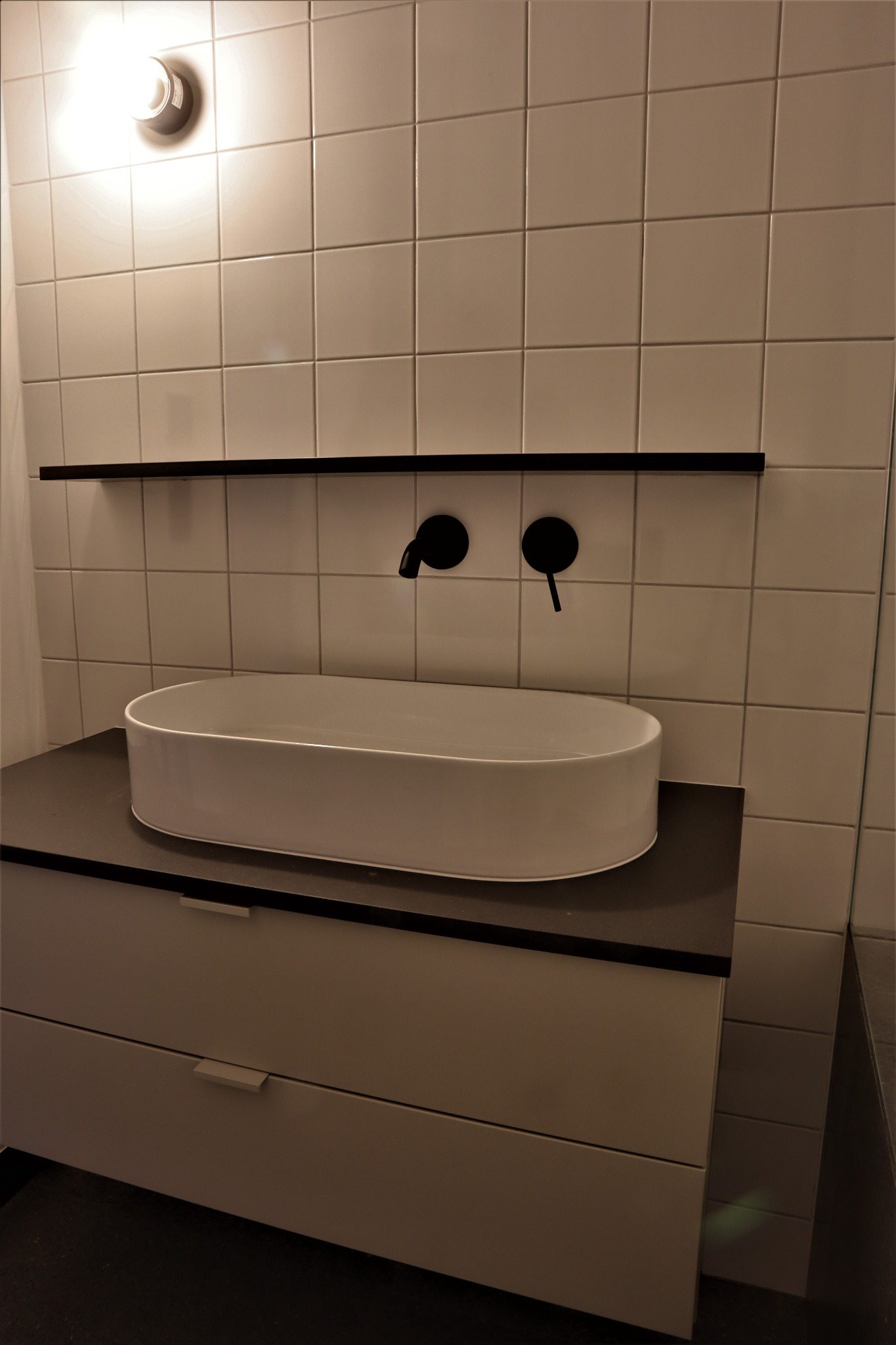 Nowoczesna łazienka z owalną umywalką nablatową, czarną armaturą i minimalistyczną szafką z szufladami, oświetlona punktowym światłem.