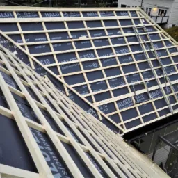 Konstrukcja dachu z łatami i kontrłatami pokryta membraną dachową z widocznymi napisami producenta, rusztowanie w tle.