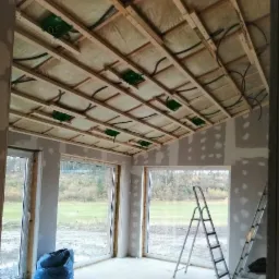 Wnętrze domu w trakcie wykańczania: ocieplony strop z widoczną konstrukcją drewnianą, instalacją elektryczną i puszkami, ściany przygotowane pod malowanie, okna z widokiem na zewnątrz, drabina...