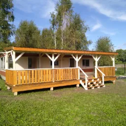 Gotowy, parterowy dom z drewnianą werandą w rustykalnym stylu, otoczony zielenią, widok z przodu, jasne niebo.