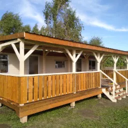 Dostawiony do domku mobilnego drewniany taras z zadaszeniem, zabezpieczony drewnianą balustradą, widok z zewnątrz w słoneczny dzień.