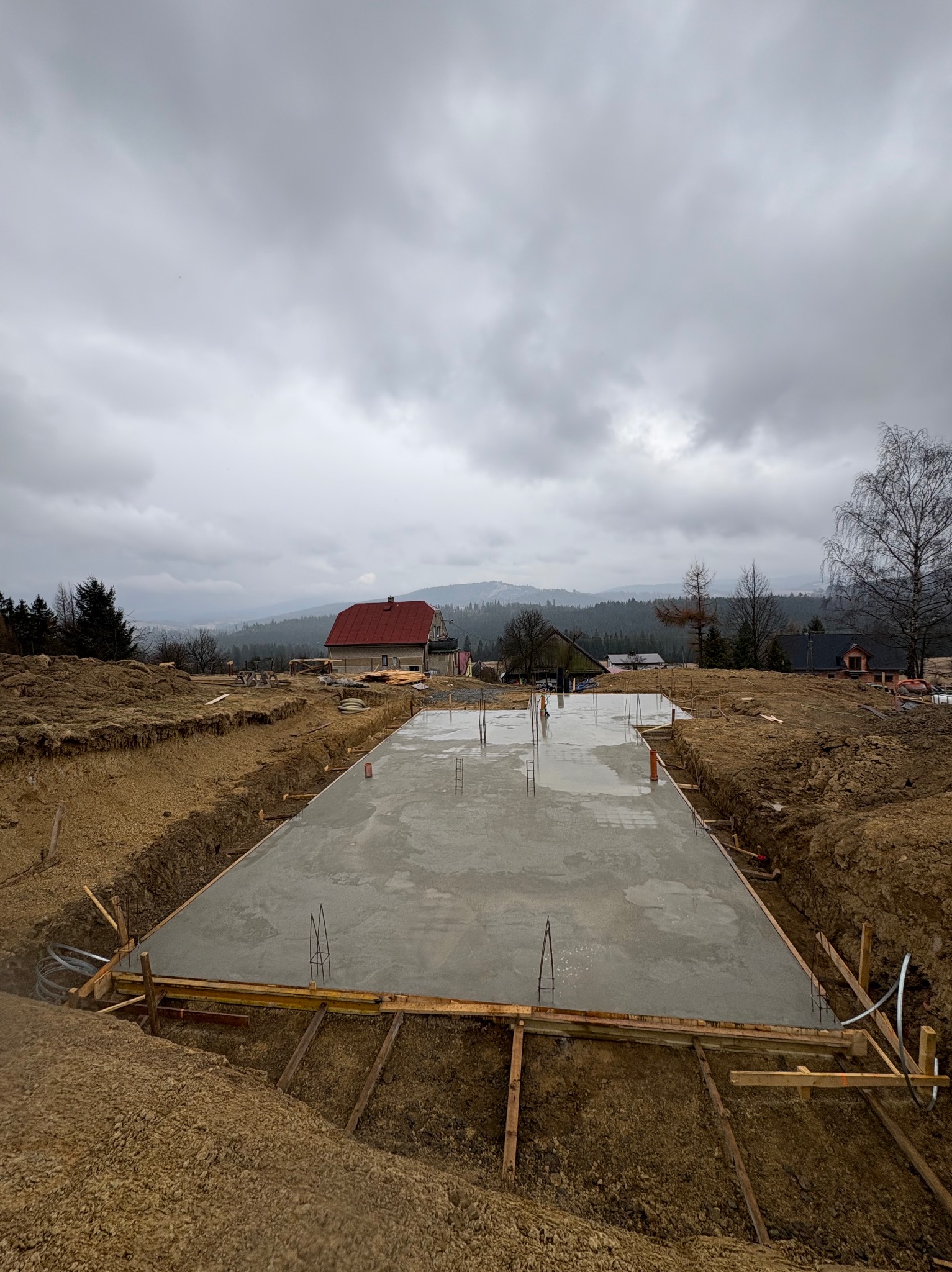 Wylana płyta fundamentowa z wystającymi prętami zbrojeniowymi, otoczona drewnianym szalunkiem na tle krajobrazu z domem z czerwoną dachówką i zalesionymi wzgórzami.