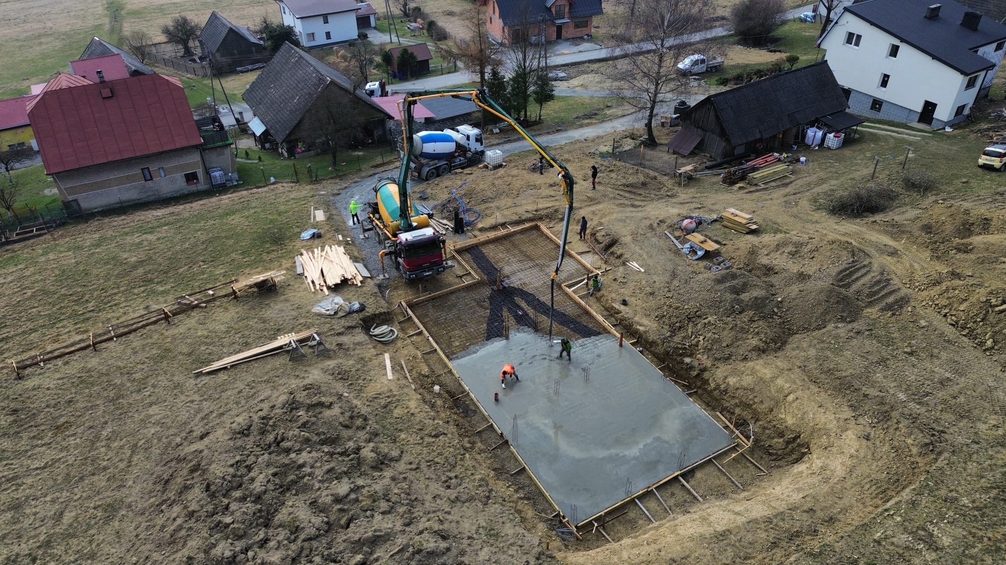 Zalewanie fundamentów pod dom jednorodzinny z użyciem pompy do betonu. Widok z góry na plac budowy w wiejskiej scenerii. W tle zabudowa mieszkalna.